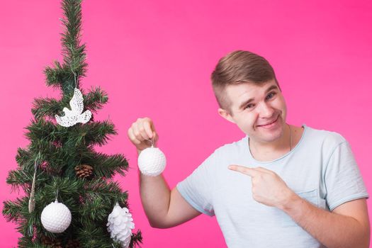 Christmas, holidays and people concept- young happy man show christmas decorations on christmas tree over the pink background.