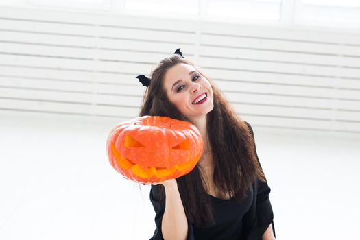 Excited happy young woman in halloween costume posing with carved pumpkin in lightroom.
