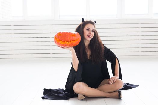 Excited happy young woman in halloween costume posing with carved pumpkin in lightroom.