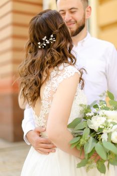 Happy smiling groom hugging bride keeping flowers and wearing white dress. Concept of married couple and love, wedding photo session.