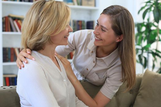 Beautiful middle aged mom and her adult daughter are hugging and smiling while sitting on couch at home