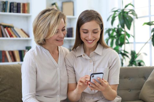 Middle aged mother and adult daughter hugging, using phone together, watching video or photos, sitting on cozy couch at home