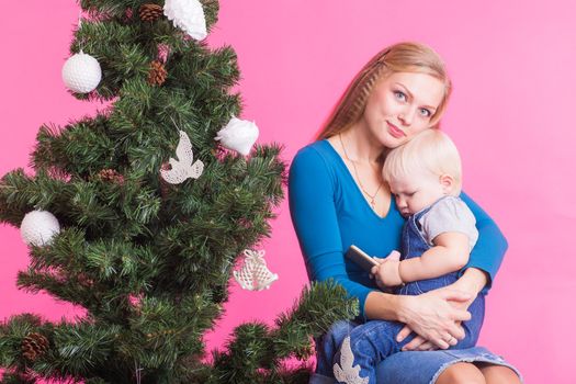 pHolidays, family and christmas concept - young woman with her baby near christmas tree on pink background.