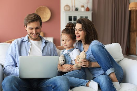 Positive friendly young parents with smiling little daughter sitting on sofa together answering video call on laptop and waving hand in greeting while relaxing at home on weekend