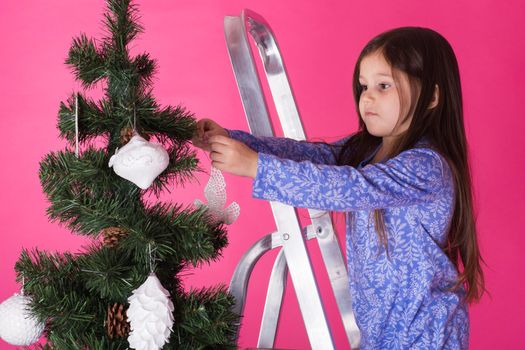 Children, holidays and christmas concept - little girl decorating christmas tree on pink background.