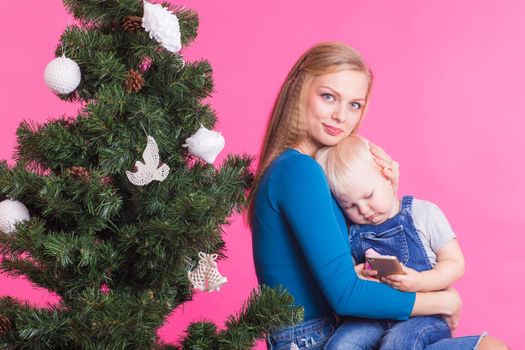 pHolidays, family and christmas concept - young woman with her baby near christmas tree on pink background.