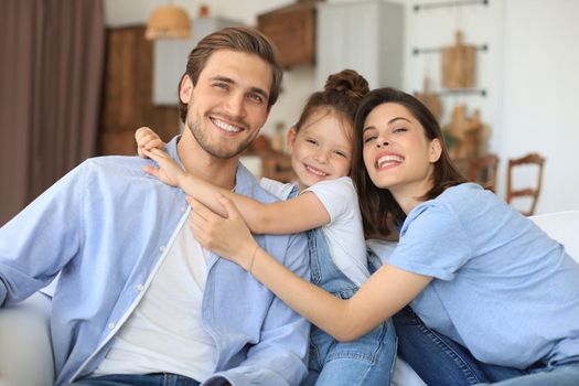 Positive friendly young parents with smiling little daughter sitting on sofa together while relaxing at home on weekend