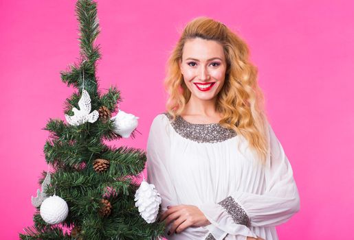 People, holidays and christmas concept - young blonde woman standing near christmas tree on pink background.