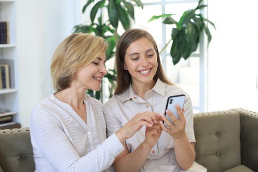 Middle aged mother and adult daughter hugging, using phone together, watching video or photos, sitting on cozy couch at home