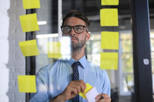 Young modern business man using adhesive notes while standing behind the glass wall in the office