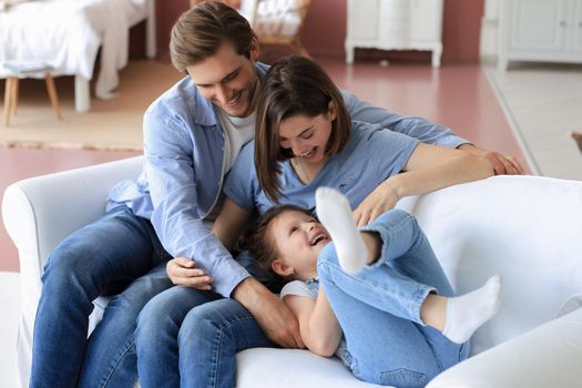 Positive friendly young parents with smiling little daughter sitting on sofa and having fun together at home
