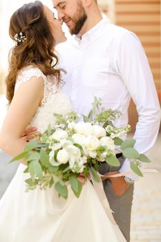 Happy american groom hugging bride keeping flowers and wearing white dress. Concept of married couple and love, wedding photo session.