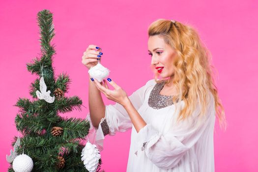 People, holidays and christmas concept - young blonde woman decorating christmas tree on pink background.