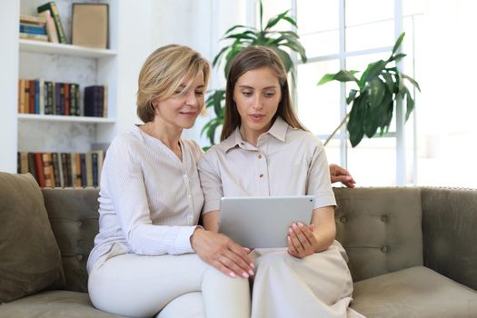 Happy mature woman with her adult daughter smiling while sitting on sofa and using laptop in living room