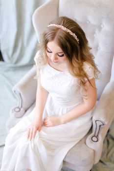 Nice fiancee sitting in arm chair at studio. Concept of bridal photo session and wedding fashion.