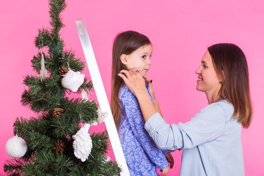 people, family and christmas concept - mother and daughter decorating christmas tree on pink background.