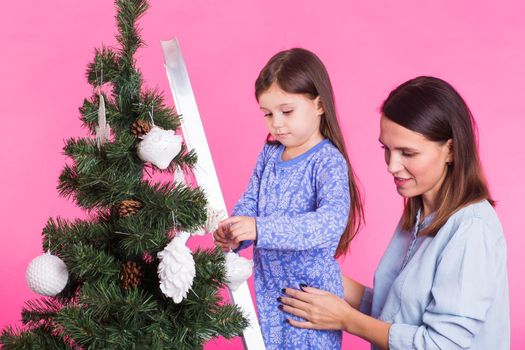 Christmas, childhood and people concept - child girl standing on step-ladder decorating the christmas tree with her mother on pink background.