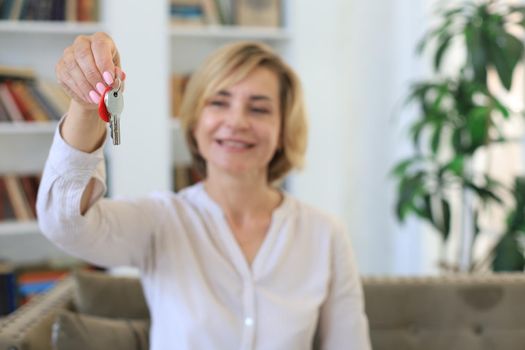 Cheerful middle aged female, real estate agent, sitting on couch, holding keys to new house