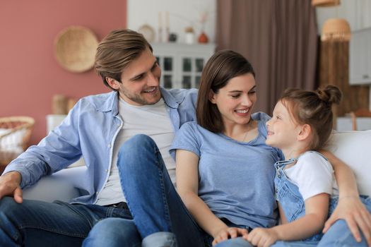 Positive friendly young parents with smiling little daughter sitting on sofa together while relaxing at home on weekend