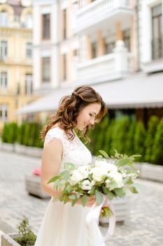 Caucasian happy fiancee walking with bouquet of flowers in city. Concept of bridal photo session.