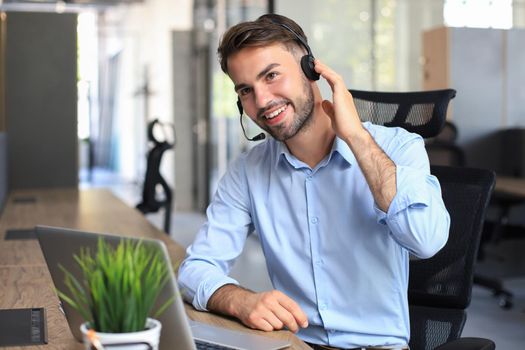 Smiling male call-center operator with headphones sitting at modern office, consulting online information in a laptop, looking up information in a file in order to be of assistance to the client