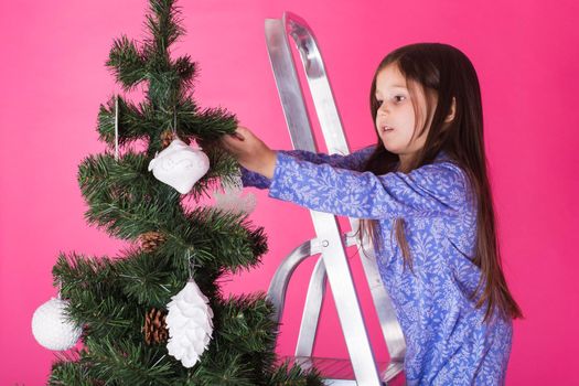 people, holidays and christmas concept - little girl decorating christmas tree on pink background.