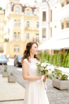 Caucasian charming fiancee walking with bouquet of flowers in city. Concept of bridal photo session.