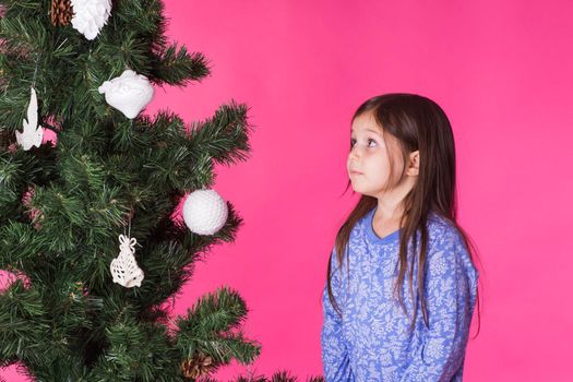 Christmas, childhood and people concept - little girl look at the christmas tree over pink background.