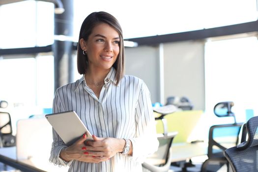 Portrait of young businesswoman looking at camera, holding touch pad while standing in modern office space interior