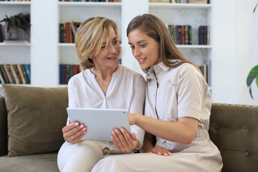 Happy mature woman with her adult daughter smiling while sitting on sofa and using digital tablet in living room