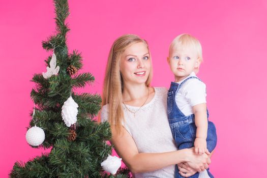 Holidays, family and christmas concept - young mom with her little daughter near christmas tree on pink background.