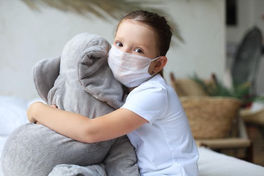 Stay at home quarantine coronavirus pandemic prevention. Sad child in protective medical mask and his elephant both in protective medical mask sit on bed