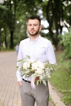 Caucasian handsome groom wearing grey trousers and white shirt waiting for bride with bouquet of flowers. Concept of wedding photo session and floristic art.