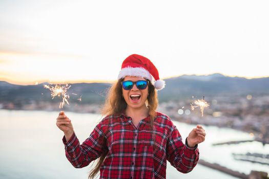 Christmas, holidays, people concept - woman in christmas hat and glasses holding sparklers over sea background.