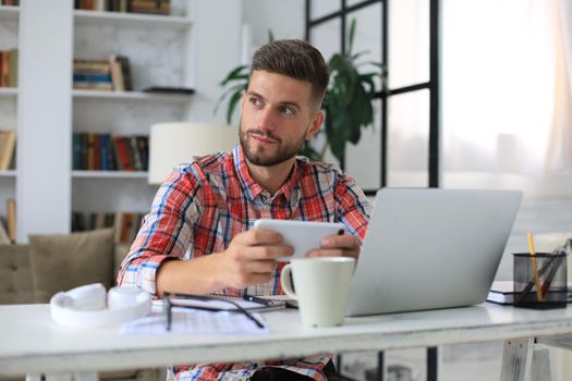 Attractive young man sitting at desk at home office and using mobile phone for cheking social nets