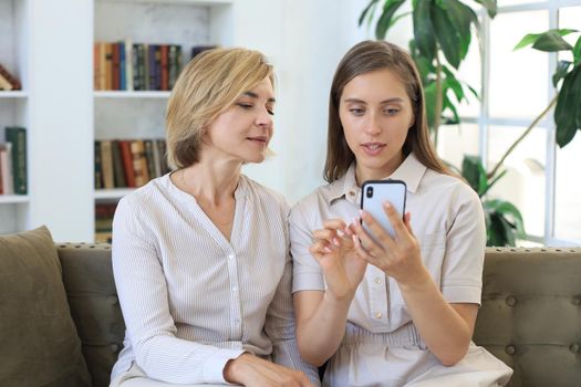 Middle aged mother and adult daughter hugging, using phone together, watching video or photos, sitting on cozy couch at home