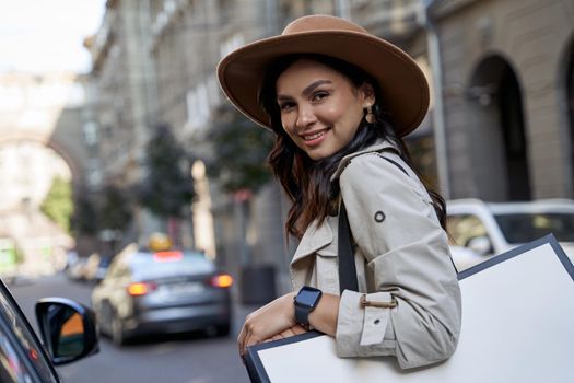 Feeling happy after shopping. Portrait of a young beautiful caucasian woman in hat with shopping bag looking at camera and smiling while standing on the city street. Fashion, people lifestyle