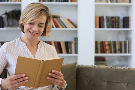 Attractive middle aged woman enjoying reading a book sitting on the sofa in her living room smiling while she reading