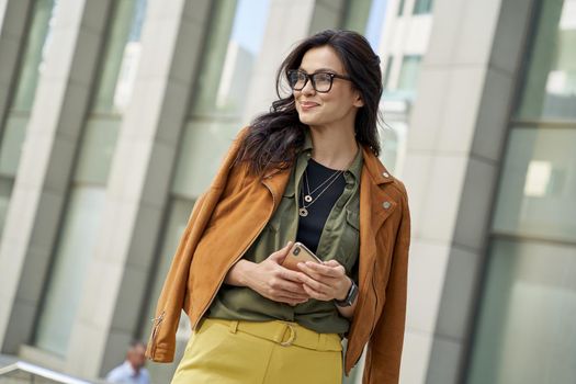 Young stylish woman holding smartphone, looking aside and smiling while standing against blurred urban background, enjoying walking on the city street. People lifestyle concept