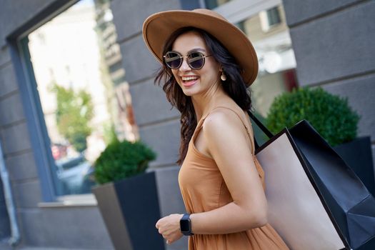 Young beautiful stylish woman wearing sunglasses, hat and summer dress with shopping bag looking at camera and smiling while standing on the city street. Fashion, people lifestyle, shopping