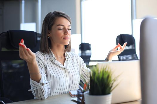 Calm beautiful businesswoman practicing yoga at work, meditating in office with eyes closed