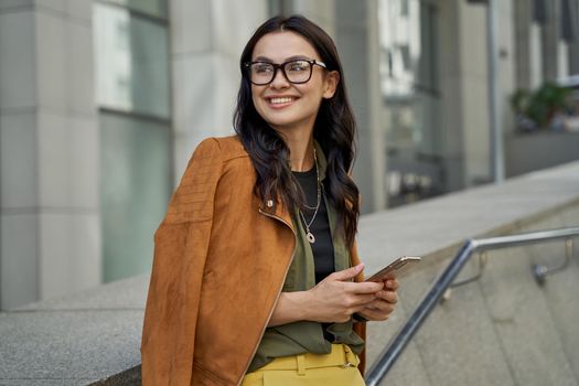 Portrait of a young beautiful and fashionable woman wearing eyeglasses holding her smartphone, looking aside and smiling while standing on the city street. People lifestyle concept