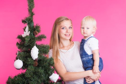 pHolidays, family and christmas concept - young woman with her baby near christmas tree on pink background.