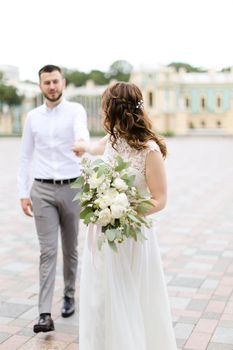 Caucasian happy fiancee walking with groom and bouquet of flowers in city. Concept of bridal photo session.