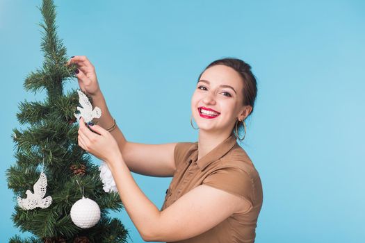 People, holidays and christmas concept - young woman decorating christmas tree over blue background.