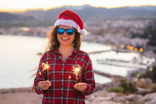 Holiday, Christmas and people concept - Young happy woman in Santa hat over beautiful landscape with fireworks and sparklers.