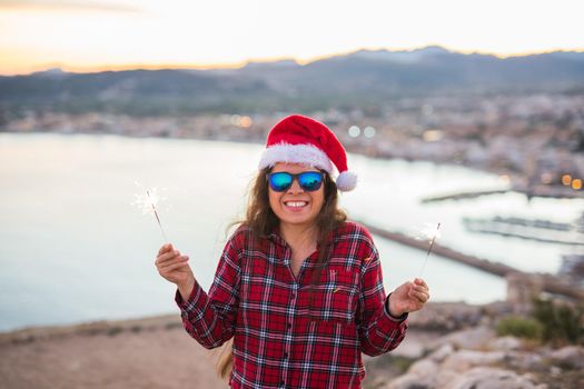 Christmas, holidays, people concept - woman in christmas hat and glasses holding sparklers over sea background.