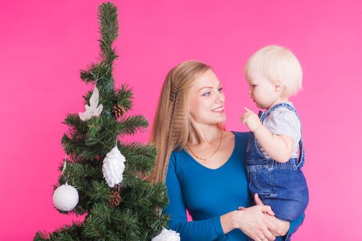 pHolidays, family and christmas concept - young woman with her baby near christmas tree on pink background.