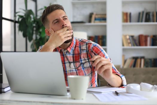 Unhappy frustrated young male holding head by hands sitting with laptop behind desk at home