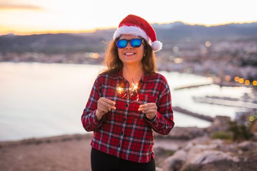 Christmas, holidays, people concept - woman in christmas hat and glasses holding sparklers over sea background.